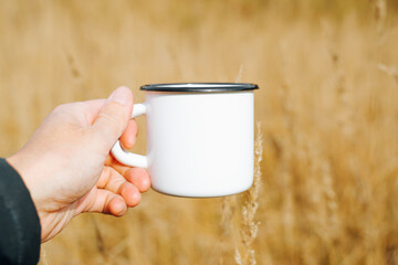 Close-up of hand showing blank white mug mockup against yellow wheat field outdoors. Enamelled...