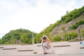 Naklejka na ściany i meble Wide shot of flexible Caucasian young yogini woman in sportswear performing yoga Pigeon pose outdoors sitting on yoga mat. Sporty calm female stretching in city park at cloudy summer day outside.