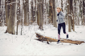 Happy fit woman jogging in woods at snowy winter day and jumping over the fallen wood. Healthy lifestyle, obstacle, success
