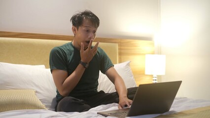 Angry Asian man is working with his laptop on his bed. Concept of freelancer problems.