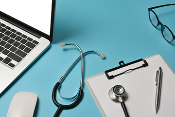 Top view of doctor's table surrounded by a white blank screen computer laptop, stethoscope, clipboard, wireless mouse, glasses and pen.
