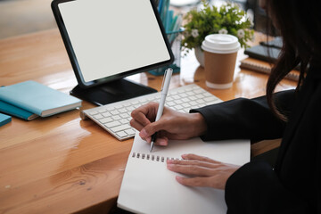 Cropped image of woman hand taking notes at the wooden table surrounded by a computer monitor, wireless keyboard, takeaway coffee cup and various equipment.