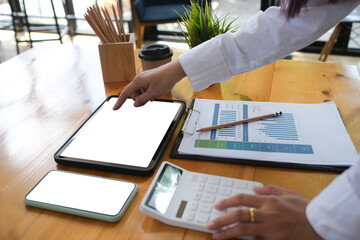 Close-up image of female finger pointing on a white blank screen digital tablet that putting on a wooden table surrounded by a white blank screen mobile, calculator and various equipment.