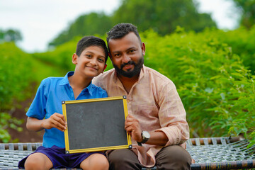 education concept : Cute indian school boy showing chalkboard with his father at agriculture field - obrazy, fototapety, plakaty