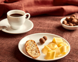 Sliced cheese, toast and sweets lie on a white plate. The plate is on a brown tablecloth. Behind the plate is a cup of coffee and a small bowl of sweets. In the background is a terracotta napkin