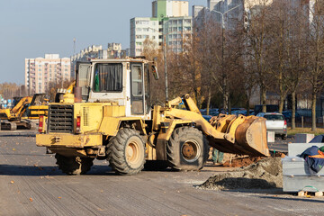 Obraz premium A heavy front loader transports earth in a bucket and grades the road. Road repair in the center of a modern city in autumn. Heavy construction machinery for moving soil.