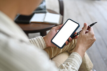 Top view image of a smart man holding a pen and using a white blank screen smartphone while sitting at the wooden vintage chair.
