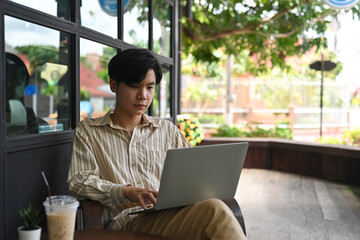 Photo of a young office man typing on a computer laptop on his lap while sitting at the wooden chair outside the restaurant as a background.