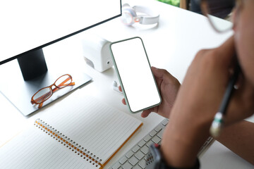 Behind view of smart man holding a white blank screen smartphone while taking notes at the working desk that surrounded by a computer monitor and various equipment.