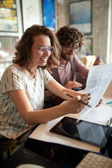 caucasian female holding paper, reading, smiling, sitting next to a beardy male colleague.