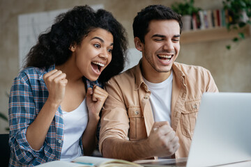 Overjoyed couple of friends using laptop, shopping online with sales, playing game looking at digital screen. Happy African American woman and Indian man win online lottery celebration success at home