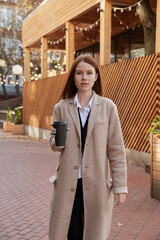 beautiful young caucasian pensive woman in beige coat with long hair standing outdoors, holding cup of coffee. attractive red headed female portrait at fall. autumn chill, lifestyle, fashion