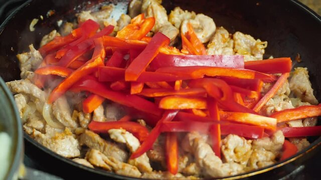 Sliced pieces of vegetables, onions, sweet peppers and meat are fried in oil and spices in cast-iron frying pan with round bottom, steam is coming, oil is boiling. Cooking stew in a cauldron, close-up