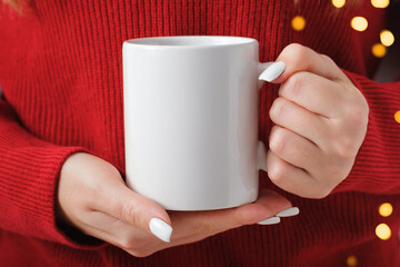 Female hands hold a mockup white tea mug in a red winter sweater, gold bokeh
