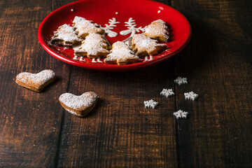 Christmas cookies in a red plate on a dark wooden table. Heart-shaped homemade baked goods sprinkled with white powdered sugar on top in a Christmas plate and signed 2022.