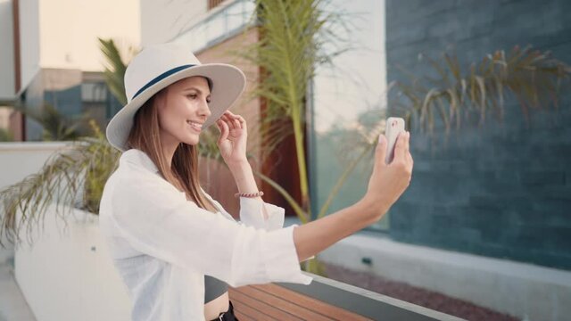 Happy young woman in stylish clothes and hat taking selfie on modern smartphone while standing on street. Female traveler saving memories from summer vacation. Happy woman taking selfie on smartphone.