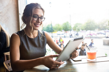 Smiling caucasian woman in glasses reading newsletter on digital gadget while sitting in coffee shop. Cheerful female student having online learning via touch pad