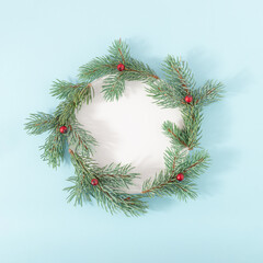 Christmas round frame made of fir branches with red berries and white paper card on bright blue background. Creative winter holiday composition. Flat lay.