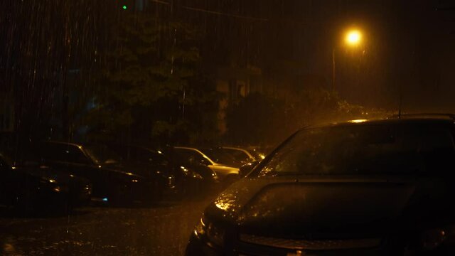 There are parked cars in a row in the courtyard of house. It's raining heavily and lightning flashes illuminating everything. Night time. strong storm hit city. Everything is flooded with water.