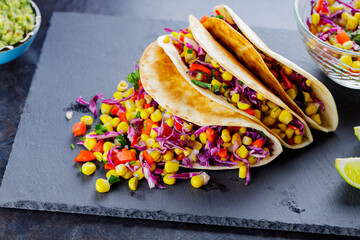 Vegetarian tacos with various vegetables, guacamole and sliced lime on dark background. Tacos with sweet corn, purple cabbage and pepper on a slate board. Copy space