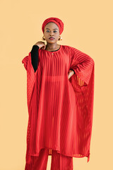 Portrait of charming pleasant African woman wearing red traditional red suit and turban, looking at camera isolated on yellow color background. Ethnic people and traditional fashion