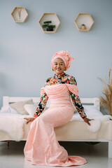 Beautiful smiling woman with bright makeup against light studio background. Studio shot of female African model in ethnic pink dress and turban, sitting on the bed, looking at camera and smiling