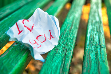 a crumpled paper note with the words I love you lies on a green bench