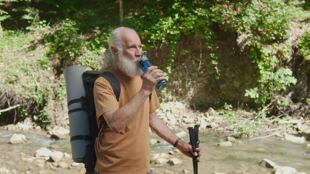 elderly man tourist drinking water from a reusable bottle on nature in the forest near the river on a sunny day. Trekking, hiking, walking. Active retirement, tourism and hobby concept.