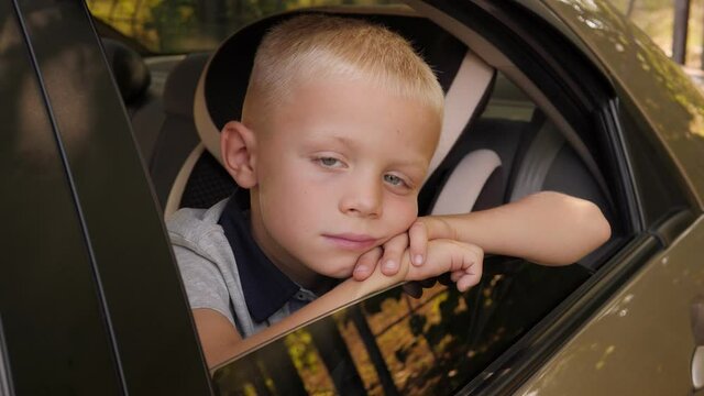 Portrait of a sad boy in the car in the back seat, he leans on the open window and looks at the camera.