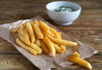 French fries lie on paper on a wooden table. Behind is a gravy boat with sauce.