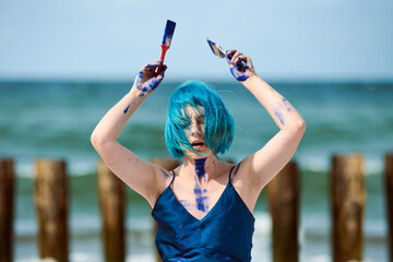 Artistic blue-haired woman performance artist smeared with blue gouache paints dancing on beach