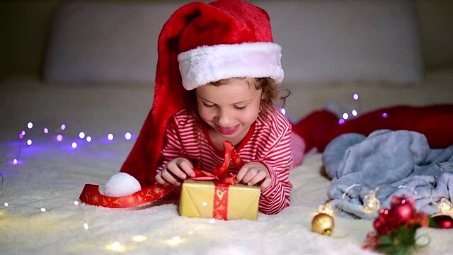 Children's Christmas. A little smiling girl in Santa's red hat and red christmas clothing lies on the bed with a gift. Preparation for the celebration. New Year. Atmosphere. Home.
