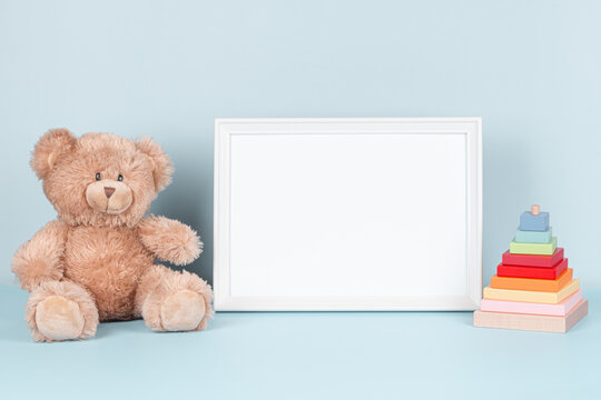 Toys background. White wooden picture frame with blank mock up copy space standing next to teddy bear and colorful wooden toy pyramid on pastel blue background. Front view