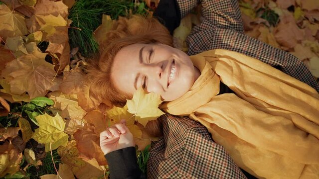 a red-haired middle-aged woman enjoys in an autumn park lying on the grass in the foliage, looking into the camera, squinting from the sun, playing with a leaf