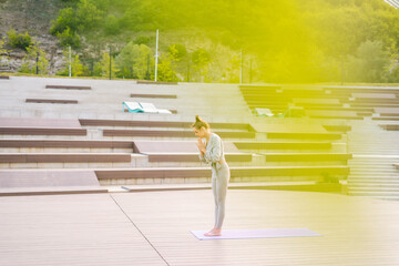 Wide shot of Caucasian young woman practicing yoga performing namaste pose with closed eyes standing on fit mat in city park. Beautiful slender female meditating and practicing yoga outdoors