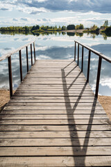 A close shoot of a wooden pier by the blue clear water, surrounded by forest on a sunny day. Solar glare on the water. Outdoor recreation by the lake. Clouds reflect in the water