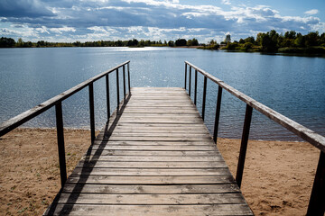 Fototapeta premium Wooden pier by the blue clear water, surrounded by forest on a sunny day. Solar glare on the water. Outdoor recreation by the lake.