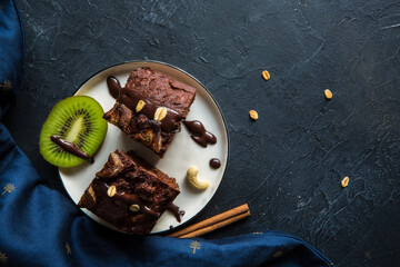 Flat lay style vegan baked brownie pies with cinnamon and chocolate on top, served on a white plate with kiwi slice on the moody dark painted background © Silent Cuisine