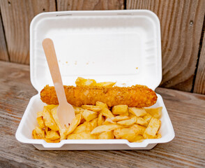 Close and selective focus on a polystyrene carton comprising a battered jumbo sausage and traditional chip shop chips cooked in beef dripping on an outdoor wooden table - obrazy, fototapety, plakaty
