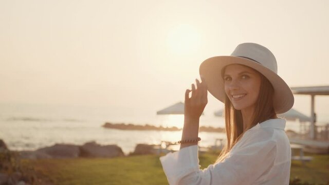 Smiling caucasian woman in white linen shirt and summer hat standing on seacoast and enjoying beautiful view. Female traveler on vacation. Lifestyles and relaxation concept.