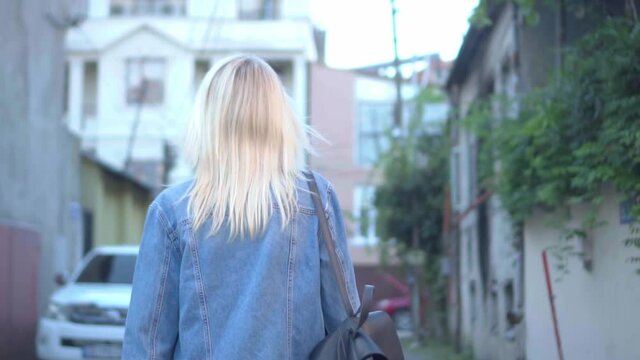 Rear view of young stylish blonde in denim jacket walking through city center along small streets of a European city. slow motion. my hair is blowing in the wind. Blurred background, out of focus.