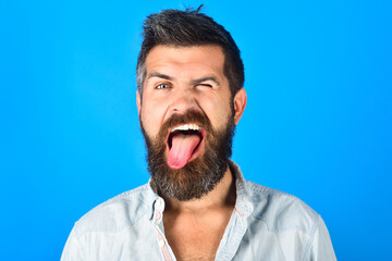 Bearded man with long beard and moustache shows tongue. Portrait of handsome bearded man on blue background. Funny faces. Emotions. Emoji.