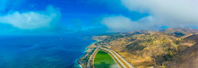 a breathtaking aerial panoramic shot of the coastline with vast blue ocean water, lush green farmland and cars on the roads along the beach with blue sky and clouds at Rincon Beach in Ventura County