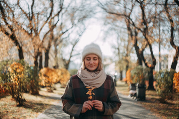 A young girl in a white hat and a plaid shirt coat stands on an autumn alley in the city and enjoys a sunny day. Holds yellow leaves in his hands