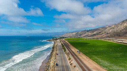 a breathtaking aerial shot of the vast blue ocean water, lush green farmland, cars on the road, majestic mountain ranges, thick cloud cover with blue sky at Rincon Beach in Ventura County California  © Marcus Jones