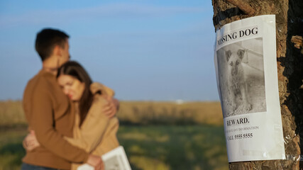 Upset couple with paper posters hugs standing in autumn park focus on report of missing dog with...