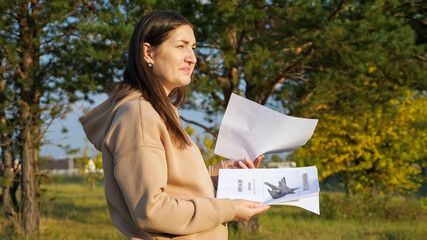 Upset young woman with long dark hair in beige hoodie looks at posters of missing cat standing in green park on sunny day side view