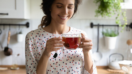 Happy woman taking tea break, holding glass mug, standing in kitchen, smelling and drinking hot...