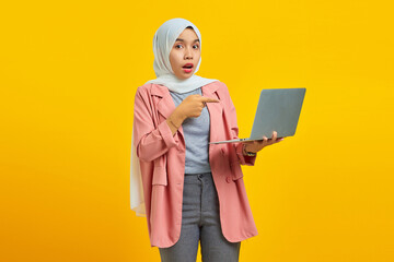 Portrait of shocked young Asian woman holding laptop and pointing isolated over yellow background