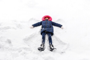 Fototapeta na wymiar Preteen happy girl in bright pink warm hood making snow angel while lying on snow outside background. Pretty child outdoor, cold weather. Caucasian kid. Fun holidays and vacations. Winter enjoy